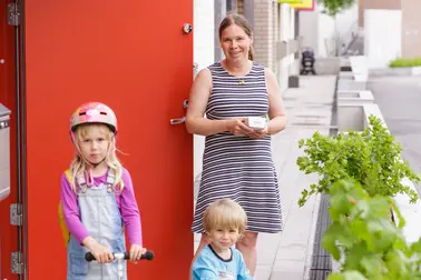 Äiti ja kaksi pientä lasta Kalasatamassa talon edessä. Tyttö on potkulaudalla ja poika pienellä pyörällä.